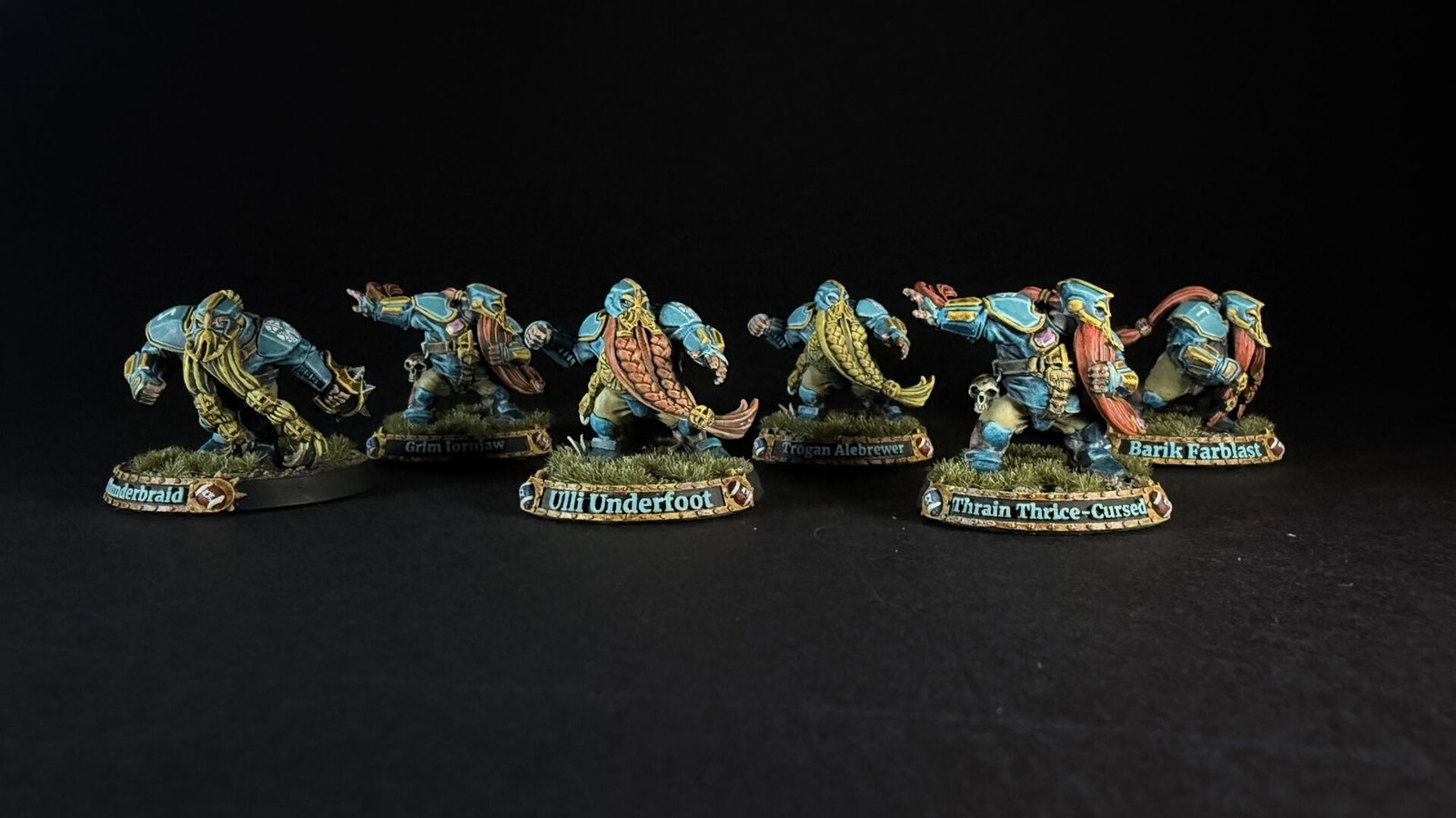 Six teal-armored dwarf miniatures with long braided beards on grassy round bases, labeled with names like Uli Underfoot and Train Thrice-Cursed.