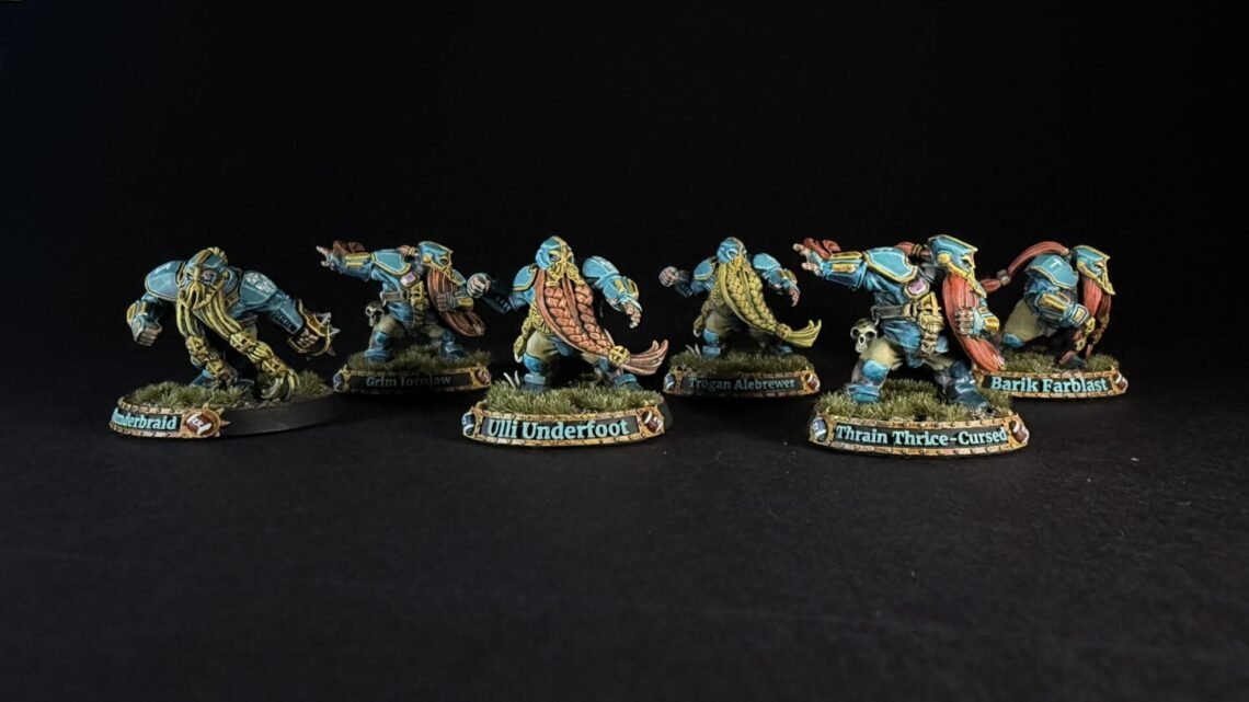 Six painted dwarf miniatures in blue armor stand in a row on grassy bases, with name banners reading Underbraid, Grim Tornjaw, Uli Underfoot, Trogan Alebrew, Train Thrice-Cursed, and Barik Farblast visible on the bases.