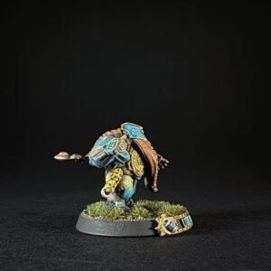 Tabletop miniature figure in blue and yellow armor, leaning forward with a brown cape on a grassy round base, set against a black backdrop (informative).