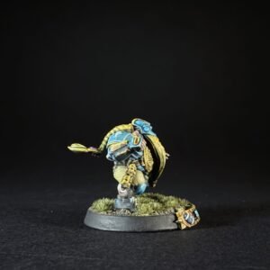 Painted miniature of a blue-and-yellow armored warrior, standing on a grassy round base with tufts, against a black backdrop.