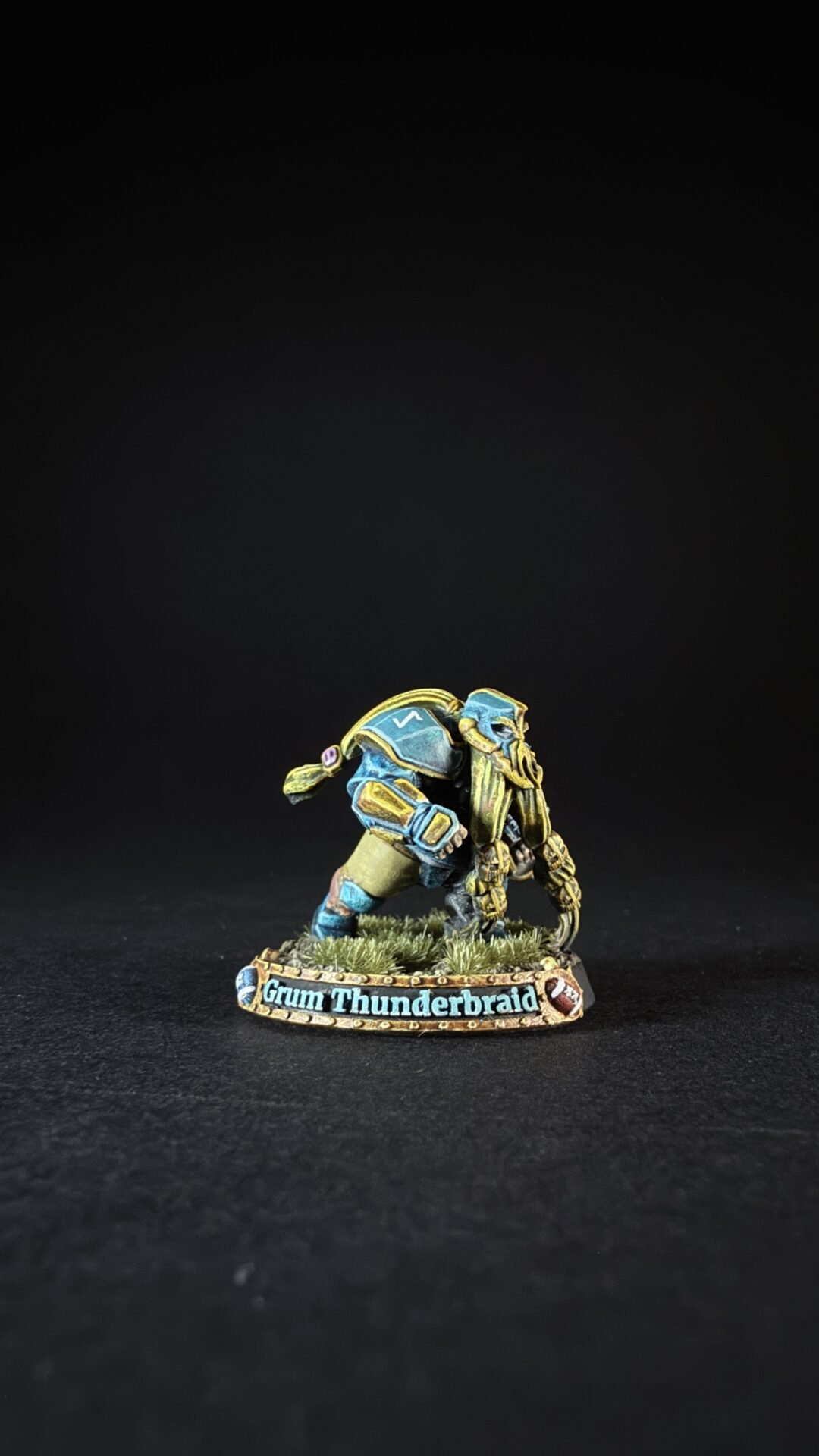 Miniature figure in blue and gold armor kneeling on grass, base labeled 'Grum Thunderbraid'.