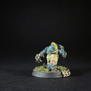 Painted fantasy miniature of a armored humanoid figure in teal and yellow, mid-stride on a grassy diorama base with a decorative emblem nearby.
