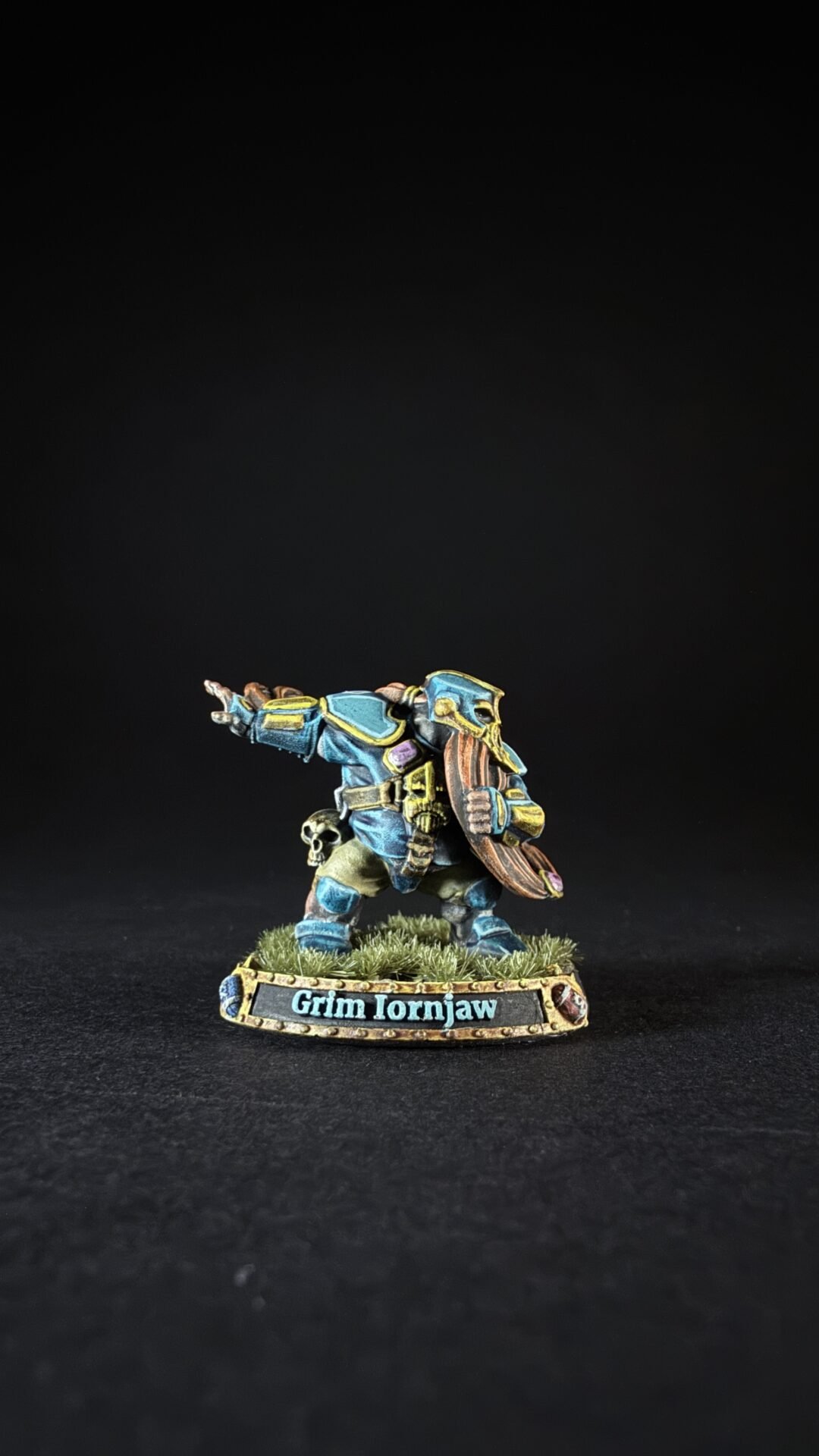 Painted dwarf warrior miniature in blue armor, pointing forward with a shield, on a grassy base labeled Grim Ironjaw.