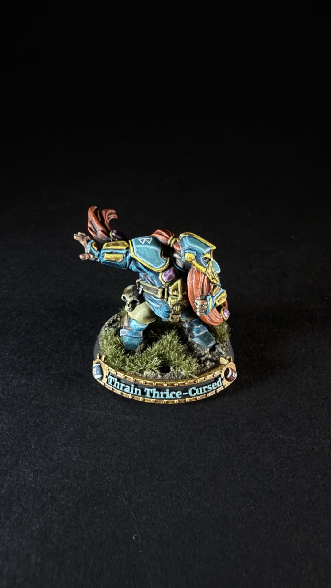 Painted dwarf warrior miniature in blue armor with an orange cape, raising a shield on a grassy rocky base labeled Thrain Thrice-Cursed.