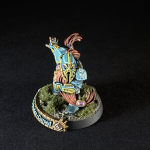 Colorful tabletop miniature of a blue-and-gold armored warrior with a raised arm, standing on a grassy base with a banner nearby.