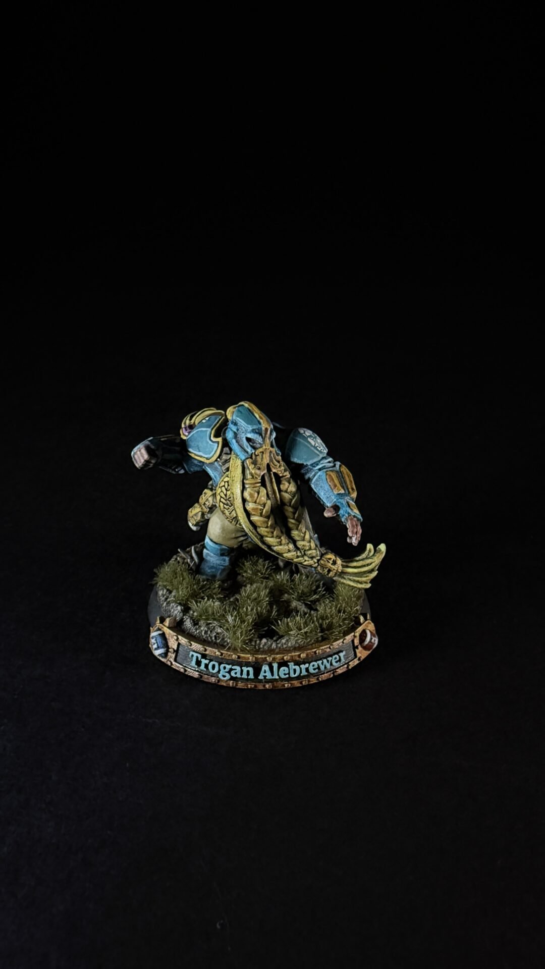 Fantasy miniature of a blue-and-gold armored fighter with a long golden cloak, standing on a grassy base; base reads 'Trogan Alebrewer'