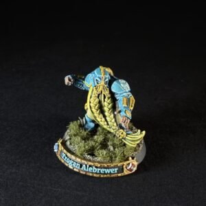 Blue-and-yellow armored miniature figure bending forward on a rocky base with pine tufts; banner reads 'Trogan Alebrew er' (text may vary due to model) and a curved base detail beneath.
