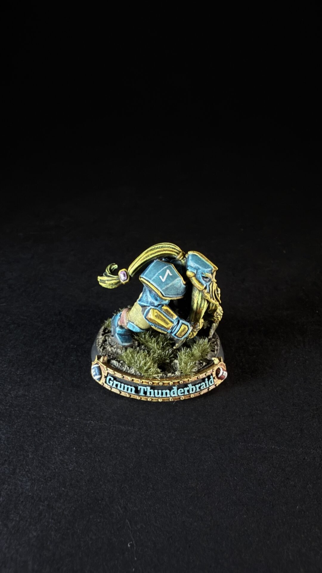 Miniature of Grum Thunderbraid dwarf in blue and gold armor, crouching on a rocky base with tufts of grass.