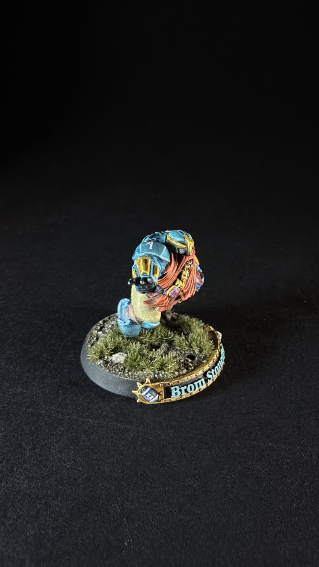 Painted fantasy miniature: blue-armored figure with orange hair on a rocky, grassy base and a decorative banner along the base.