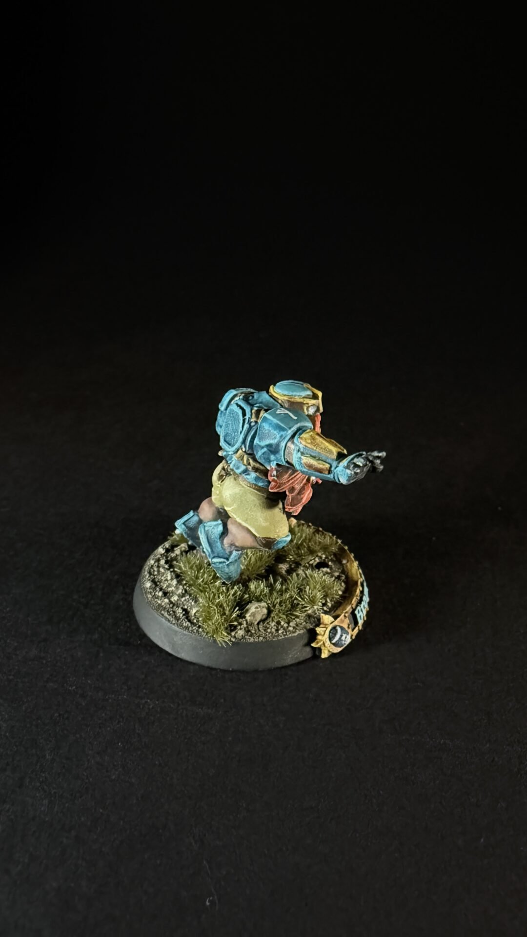 Painted tabletop miniature of a blue-and-gold armored sci‑fi soldier charging forward on a grassy base.