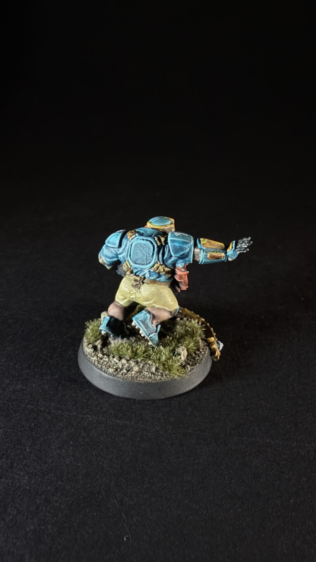 Back view of a blue armored miniature figure running on a grassy, rocky base