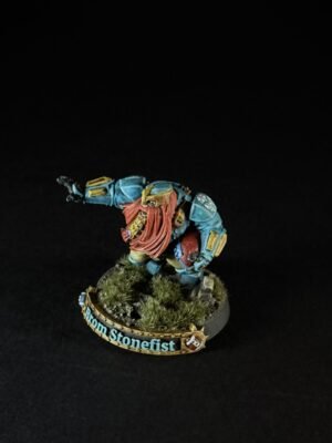 Tabletop miniature of a blue-armored dwarf with a red beard, posed as if charging forward on a rocky base with shrubs; base label reads 'Bron Stonefist'.