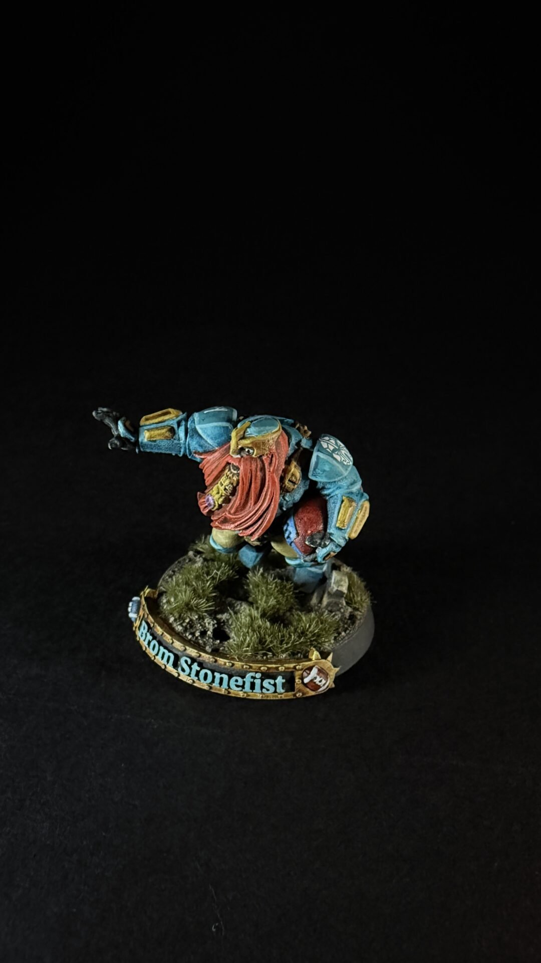 Tabletop miniature of a blue-armored dwarf with a red beard, posed as if charging forward on a rocky base with shrubs; base label reads 'Bron Stonefist'.