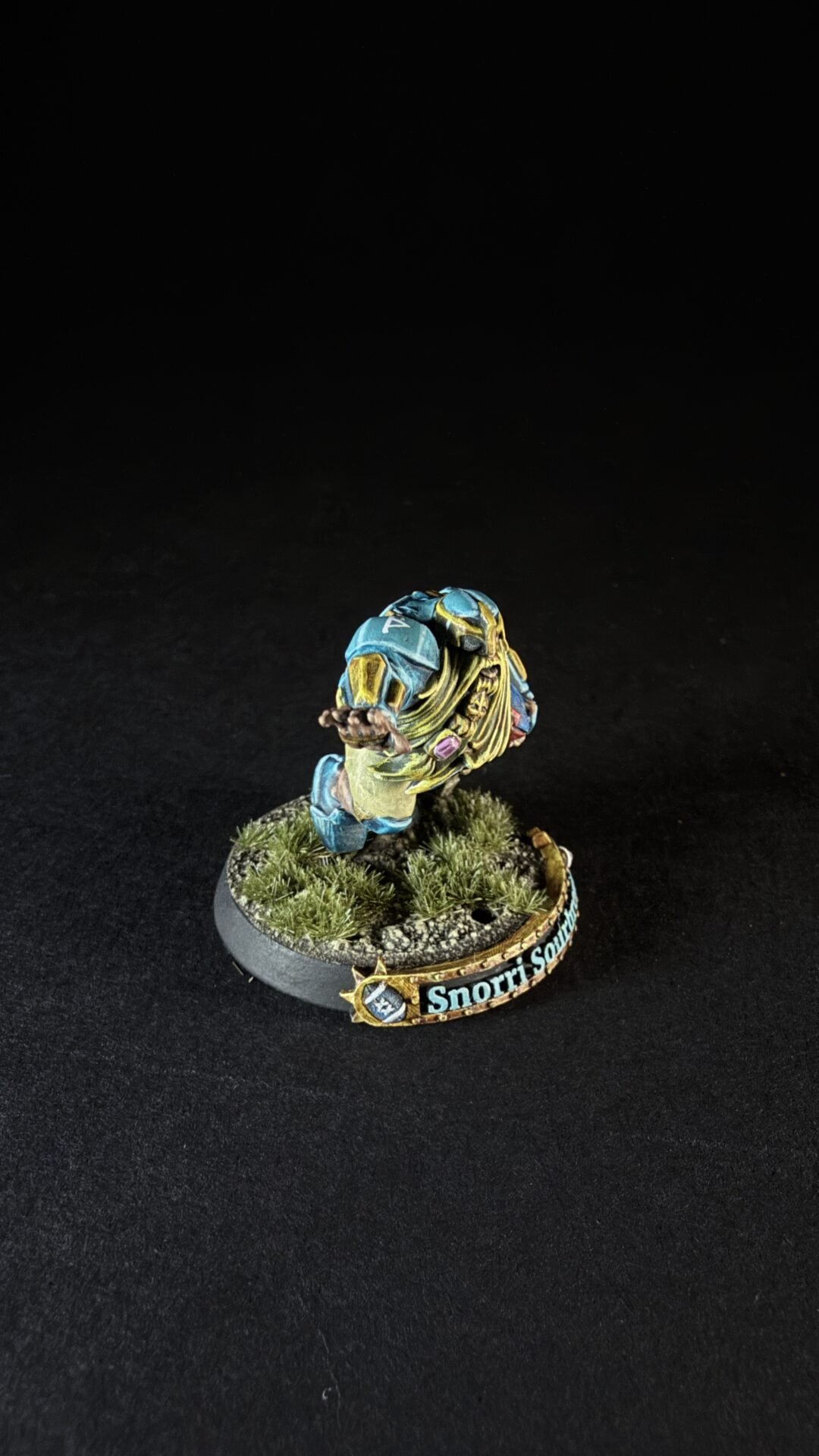 Painted miniature of a blue-and-yellow armored figure leaning forward with one arm extended, standing on a grassy diorama base labeled 'Snorri S...'