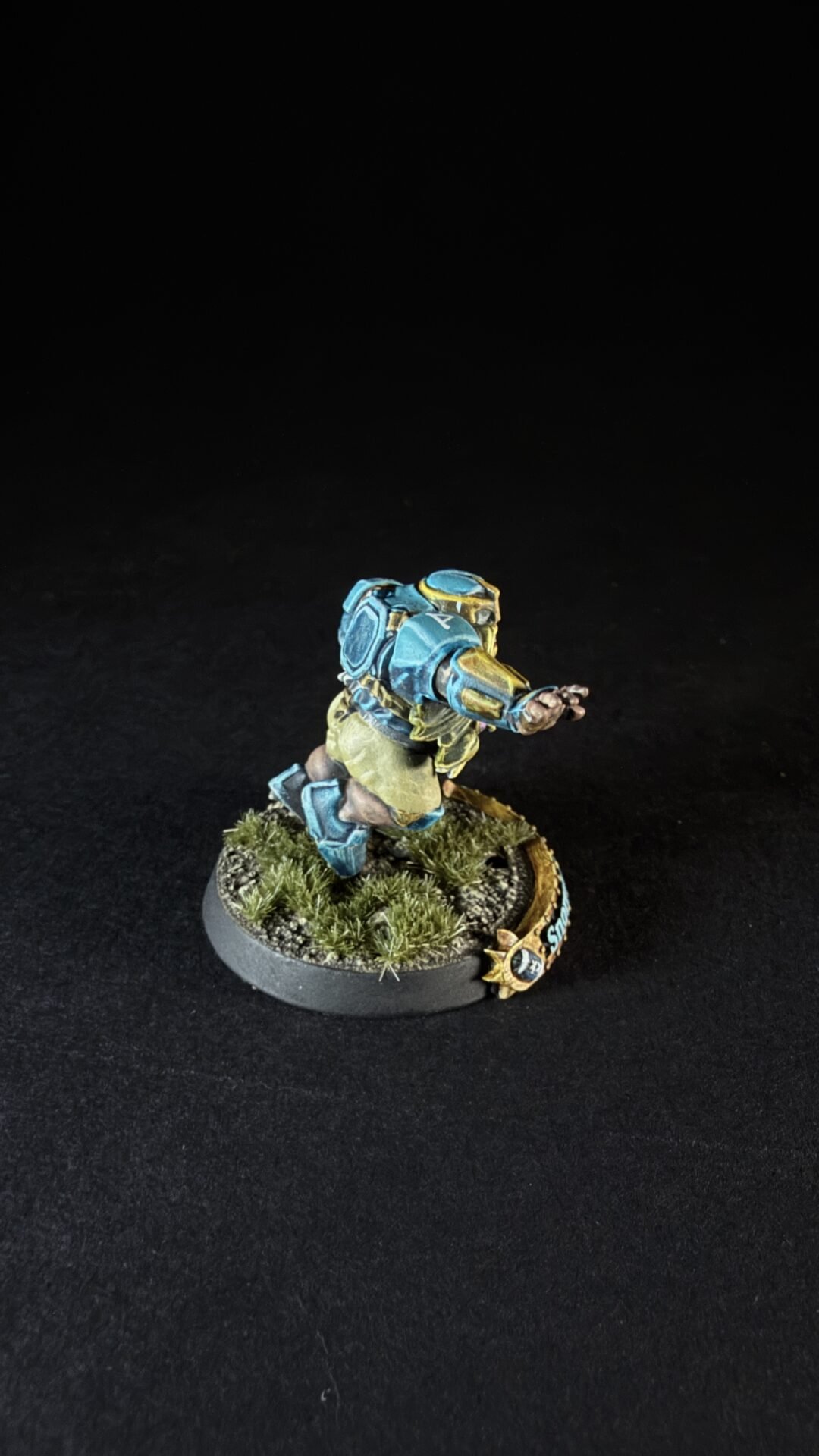 Miniature sci-fi soldier in blue armor sprinting forward, backpack pack and desert-style base with grass tufts on a round base.