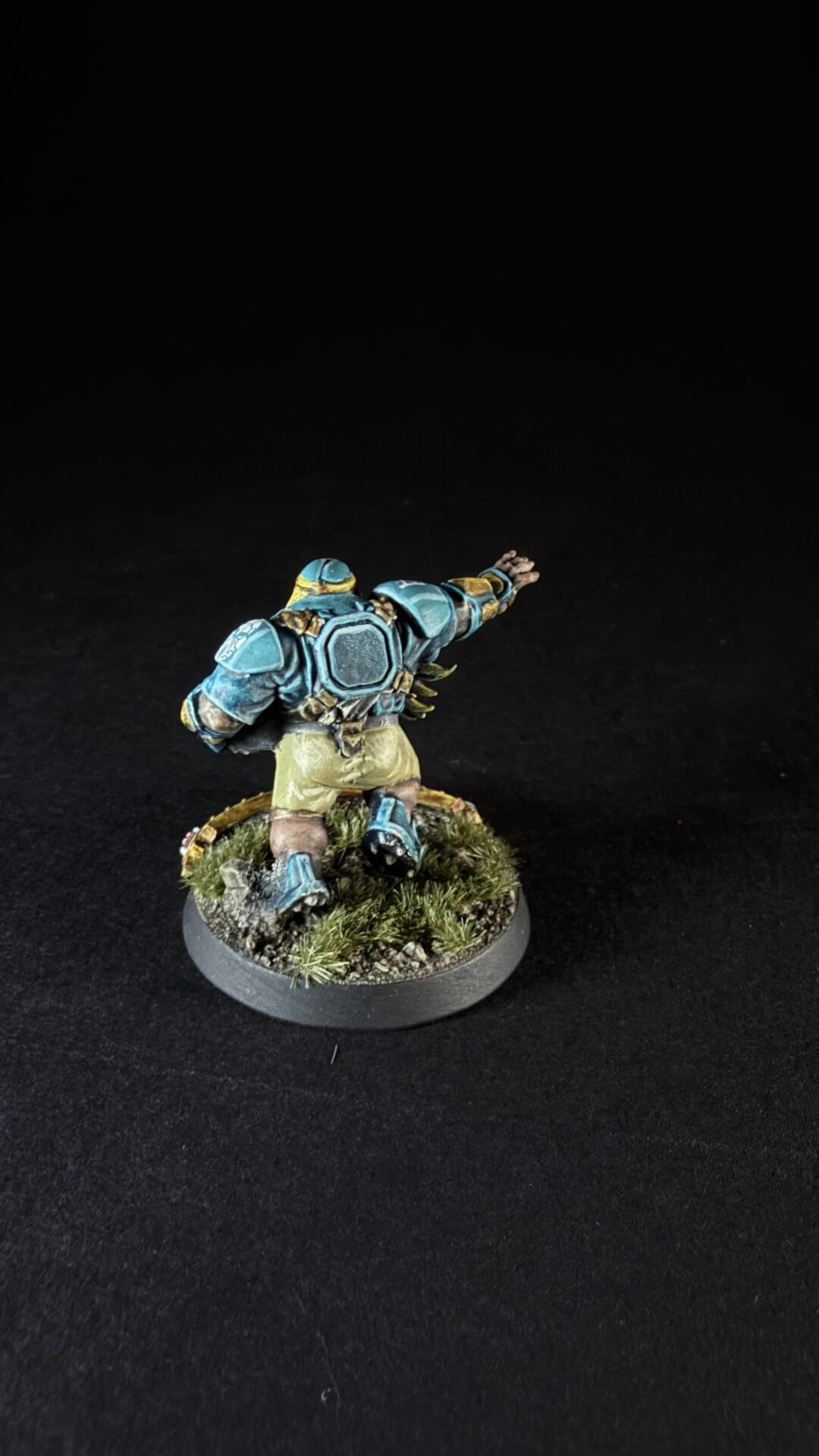 Miniature sci‑fi armored figure seen from behind, blue armor, outstretched right arm, standing on grassy battlefield base.