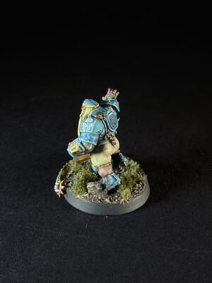 Small fantasy dwarf miniature in blue armor with yellow accents, standing on a grassy rocky base with one arm raised.