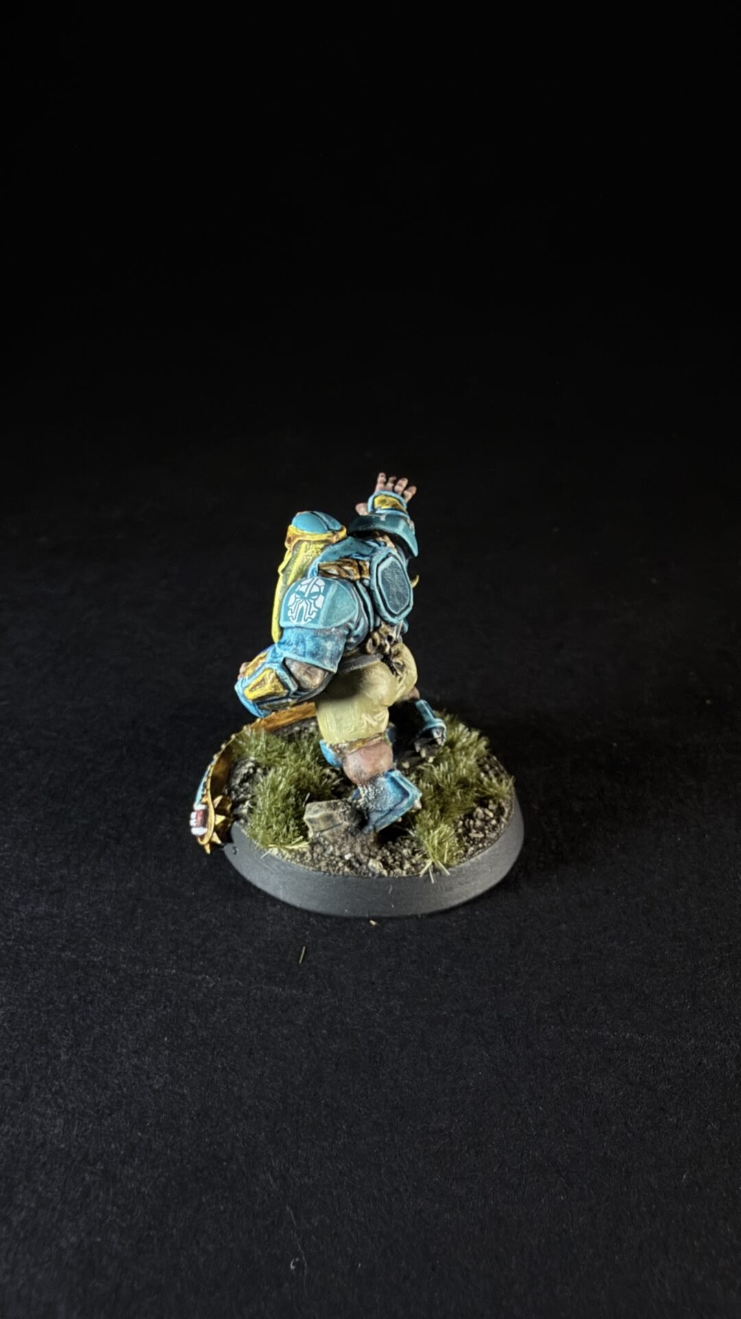 Small fantasy dwarf miniature in blue armor with yellow accents, standing on a grassy rocky base with one arm raised.