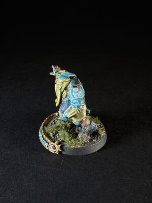 Painted sci‑fi armored miniature in blue and yellow, posed as if running, on a grassy round diorama base with a decorative banner and emblem nearby.