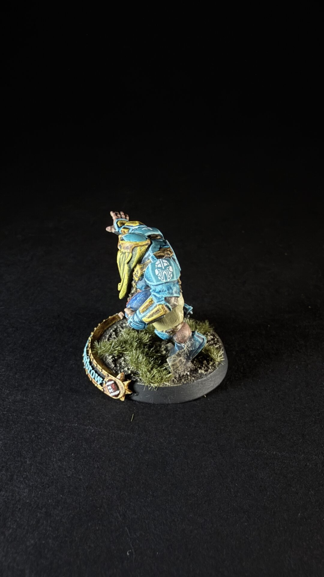 Painted sci‑fi armored miniature in blue and yellow, posed as if running, on a grassy round diorama base with a decorative banner and emblem nearby.