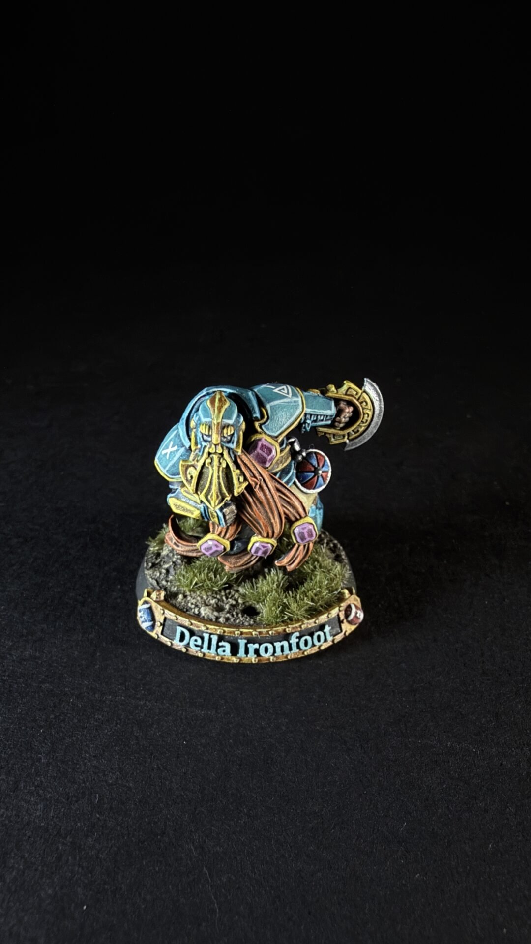Painted dwarf miniature named Della Ironfoot, wearing blue armor and a golden helm, wielding a large axe on a rocky base with tufts of grass.