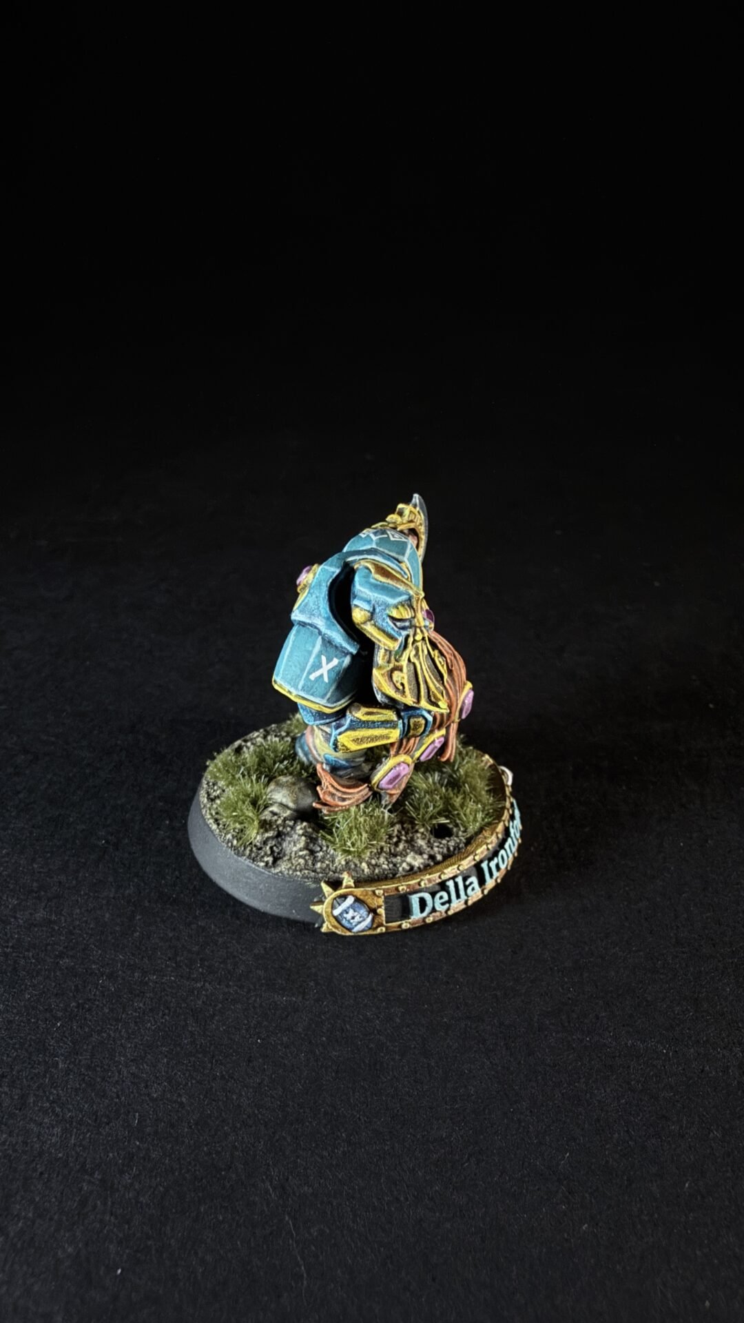 Colorfully painted fantasy miniature: a blue-armored knight with gold trim on a grassy, rocky base, name plaque reading 'Della Iron' (rear view).