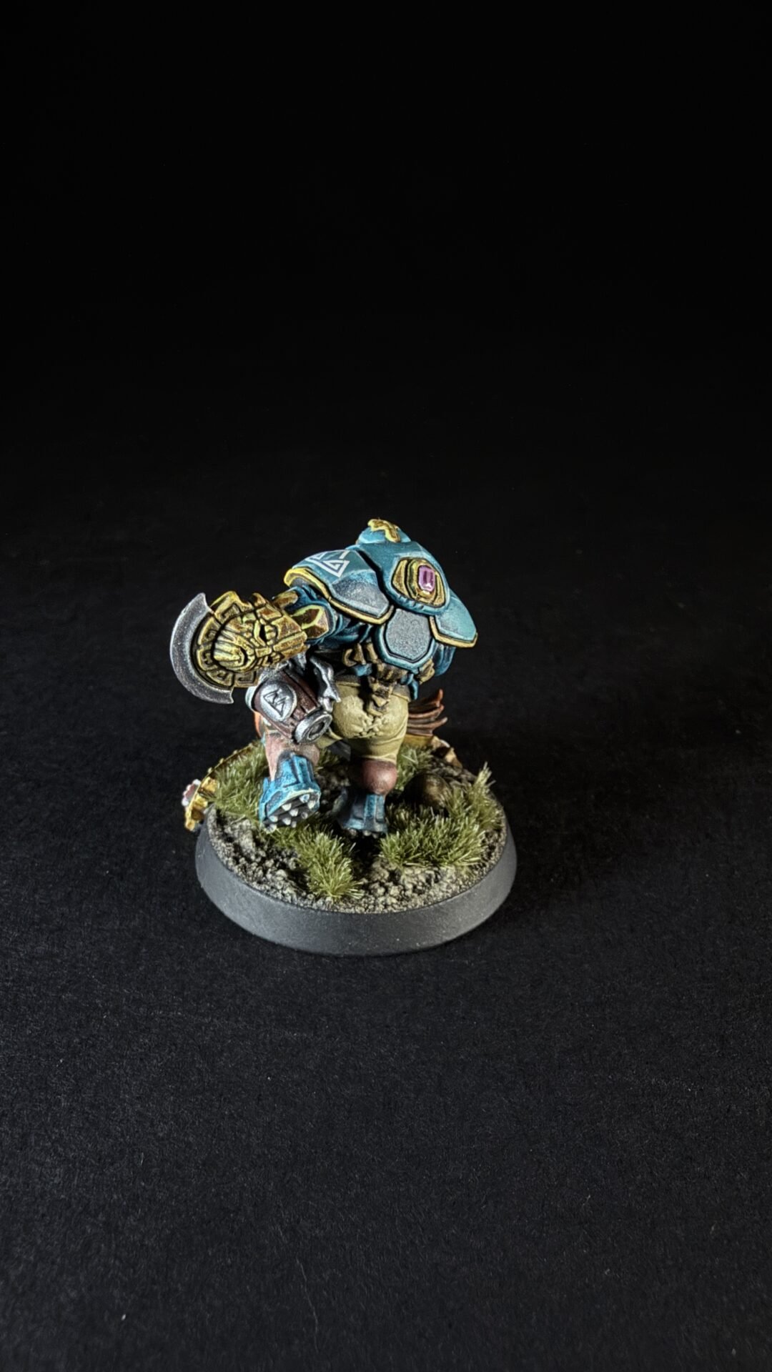 Painted fantasy dwarf warrior miniature in teal armor with gold trim, holding a large hammer, on a grassy round base.