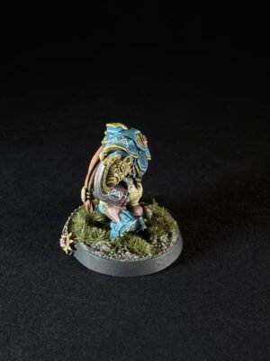 Small painted miniature figure in blue armor crouching on a grassy base with a backpack, viewed from the side.