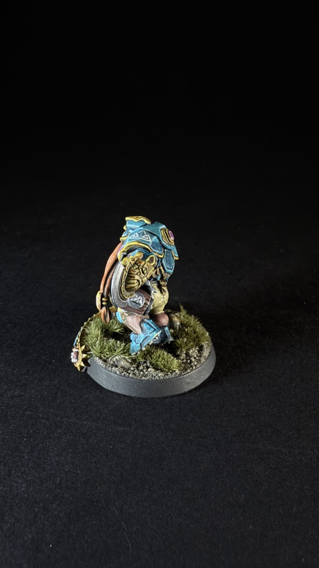 Small painted miniature figure in blue armor crouching on a grassy base with a backpack, viewed from the side.