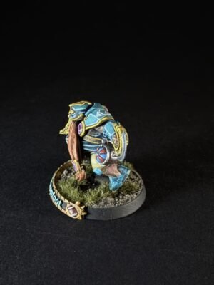Painted blue-armored tabletop miniature figure with yellow trim, standing on a grassy diorama base on a black background.