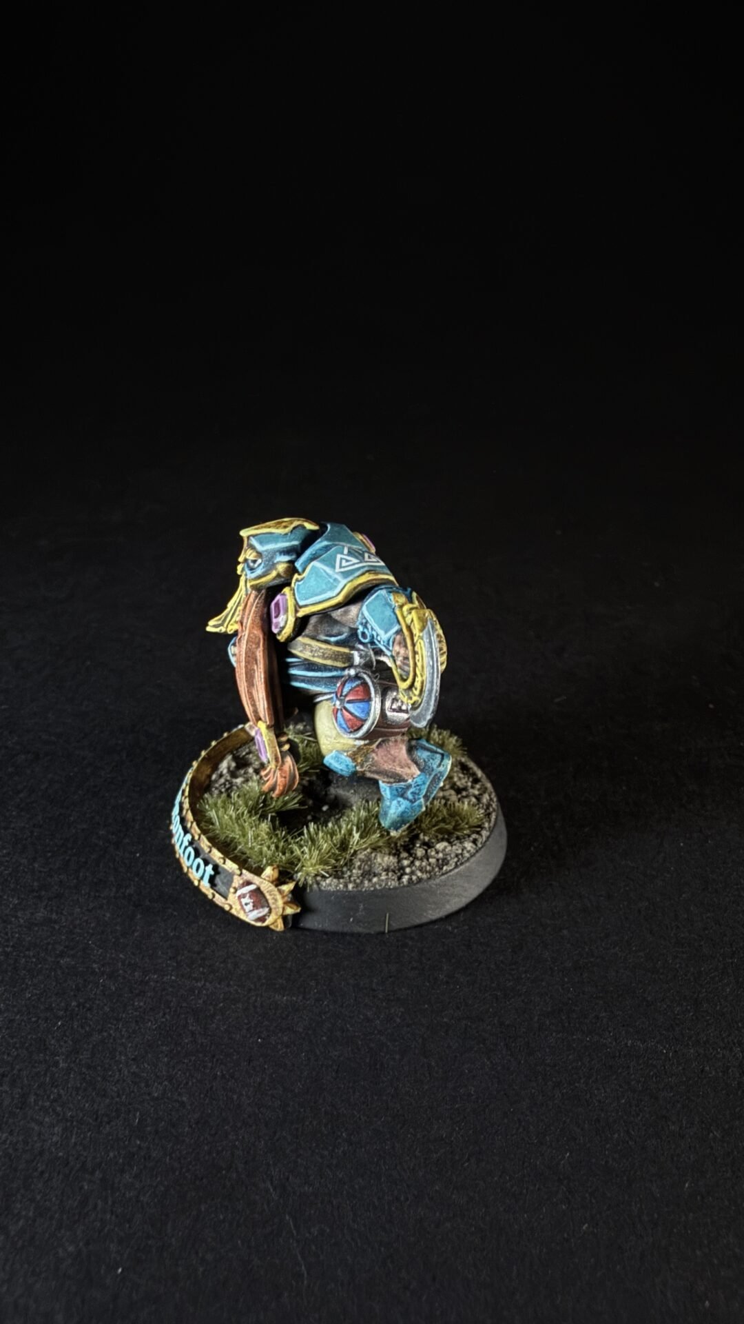 Painted blue-armored tabletop miniature figure with yellow trim, standing on a grassy diorama base on a black background.