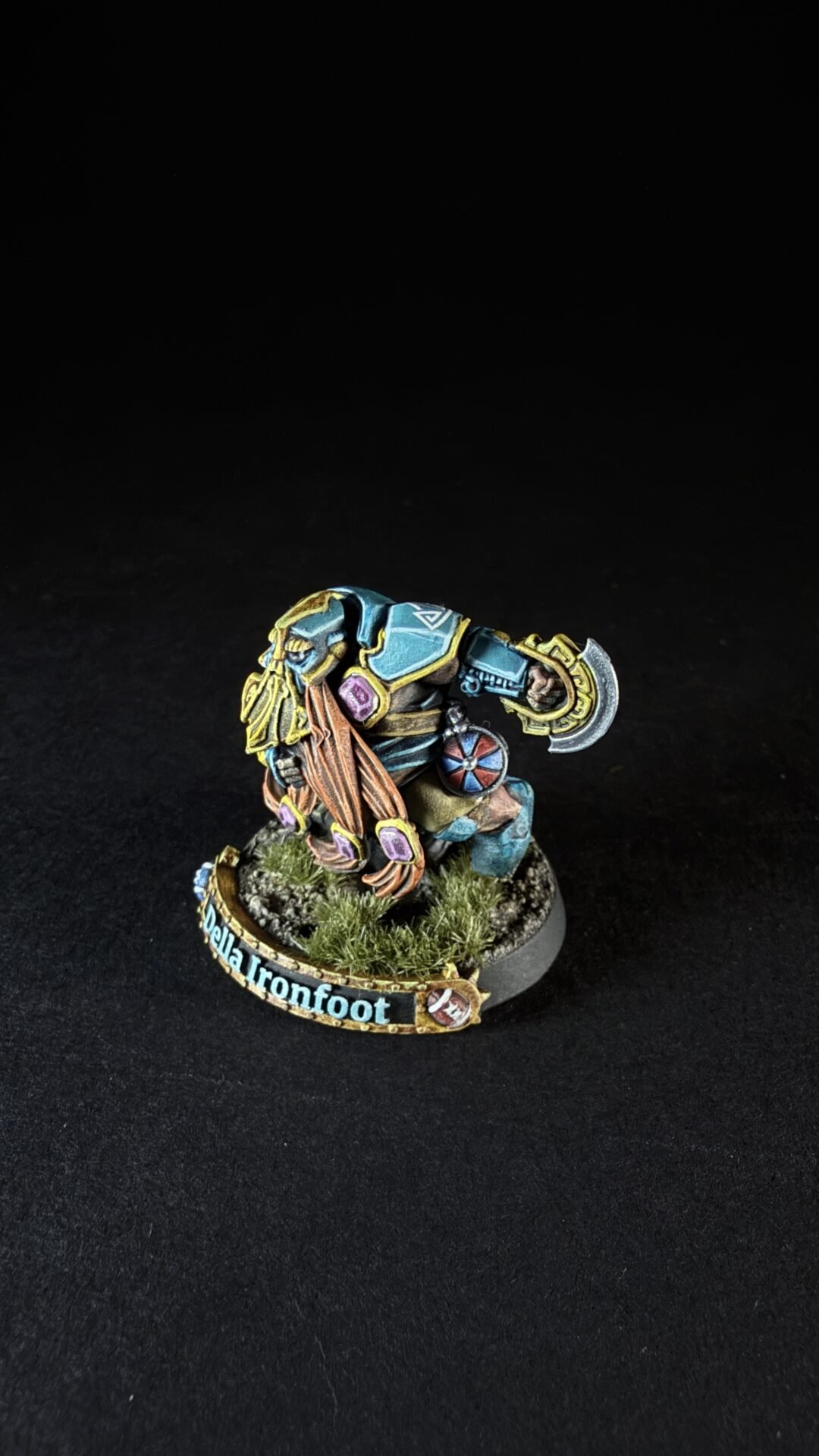 Painted dwarf miniature named Della Ironfoot, in teal armor, wielding an axe and shield, on a grassy base with tufts of grass.