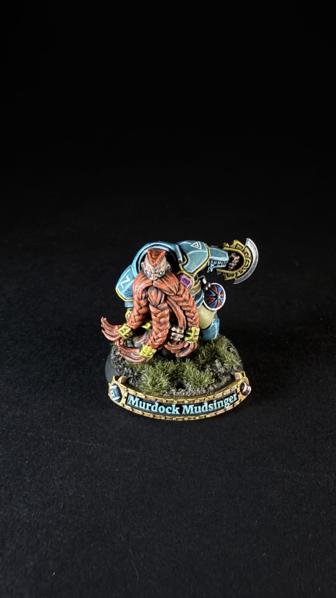 Fantasy tabletop miniature of a dwarf with a red braided beard, blue armor, and a large hammer, on a grassy base labeled “Murdock Mudsinger.”