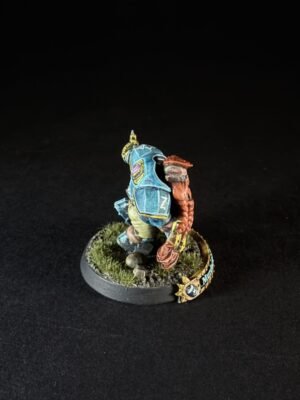 Back view of a painted tabletop miniatur e figure in blue armor with a brown braid or rope, standing on a grassy, rocky base on a black background.