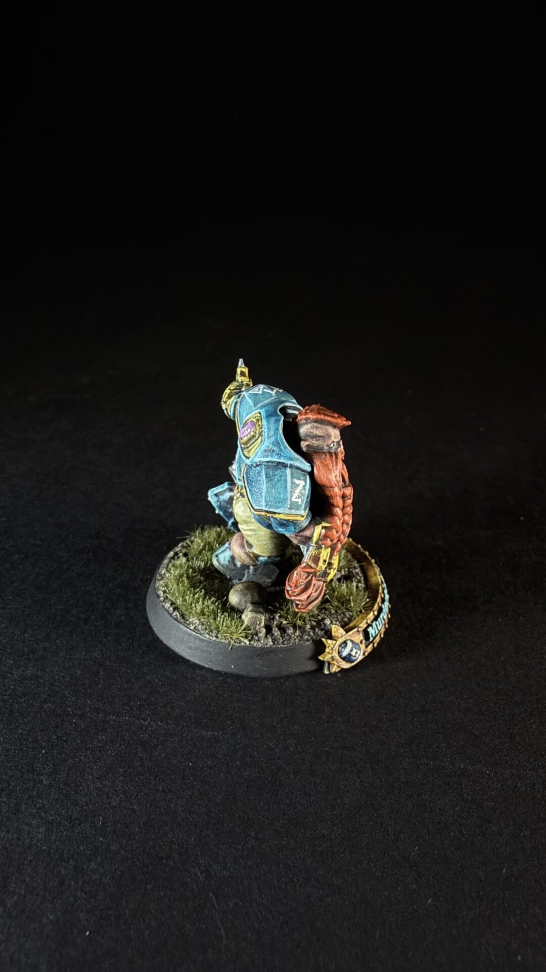 Back view of a painted tabletop miniatur e figure in blue armor with a brown braid or rope, standing on a grassy, rocky base on a black background.