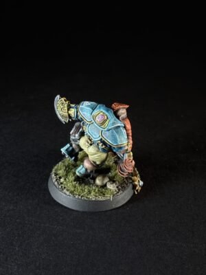 Miniature figure of a fantasy warrior wearing blue armored plates with gold trim, standing on a grassy rocky base.
