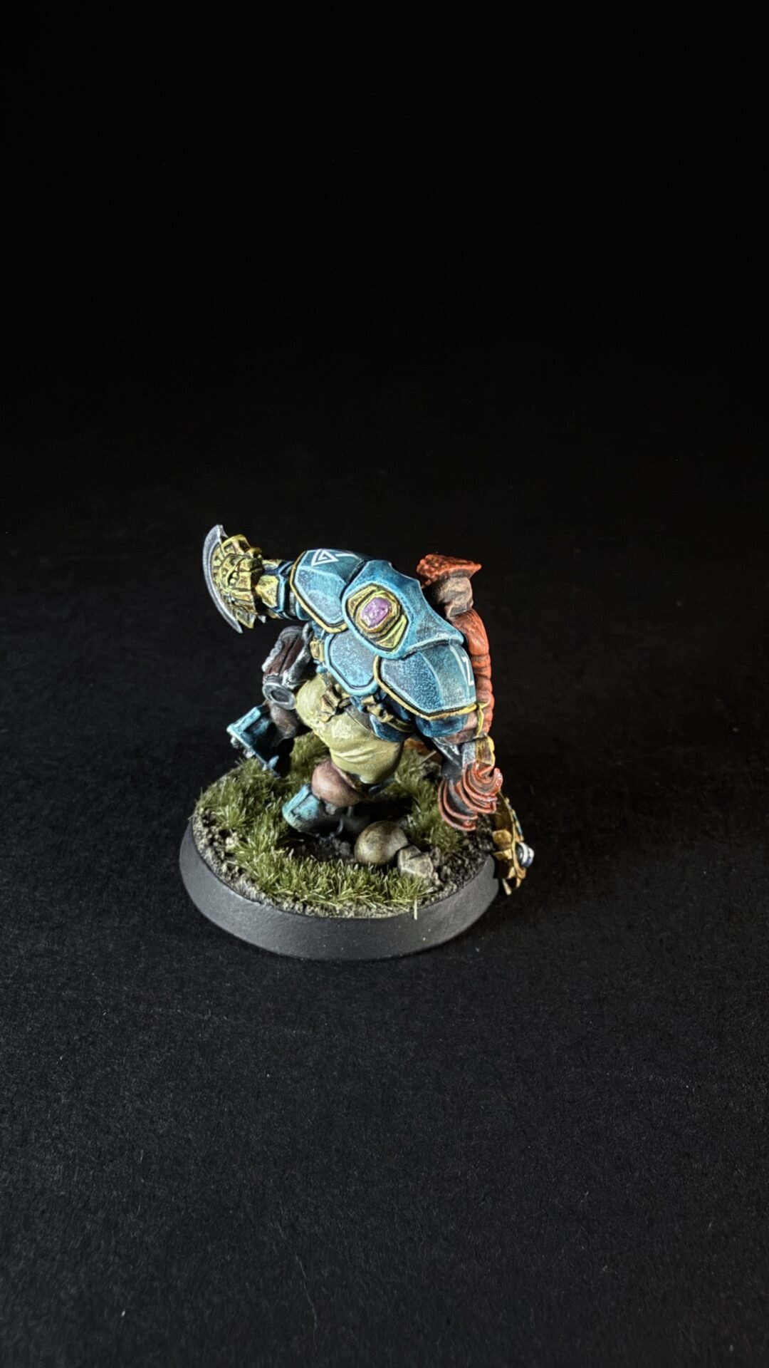 Miniature figure of a fantasy warrior wearing blue armored plates with gold trim, standing on a grassy rocky base.
