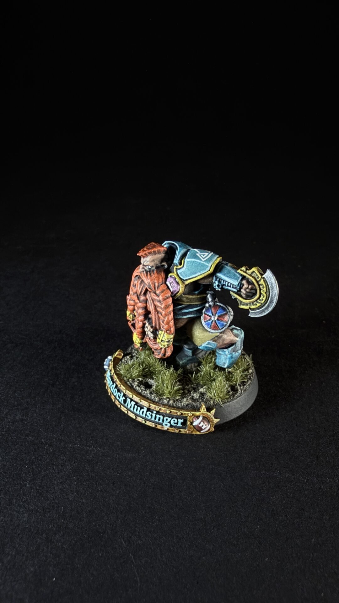 Miniature dwarf warrior in teal-blue cloak and orange beard, wielding a large axe and shield, posed on a grassy base with a curved name banner reading 'Mudstinger'.