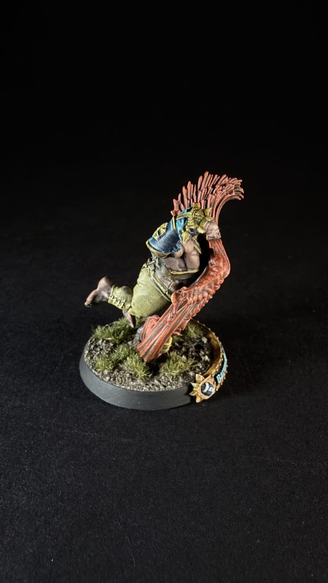 Painted fantasy miniature of a green orc-like warrior leaping with a large red spiked crest or wing behind him, on a rocky base with tufts of grass.