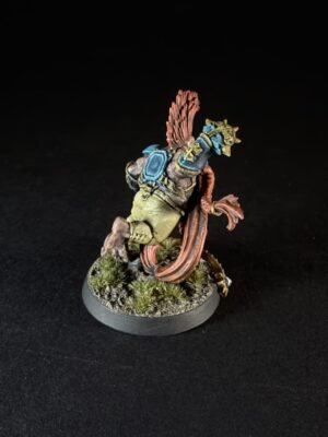 Miniature figure of a warrior with blue armor, beige clothing, and red feathered wings, leaning forward on a textured base with grass and rocks, seen from behind-right.