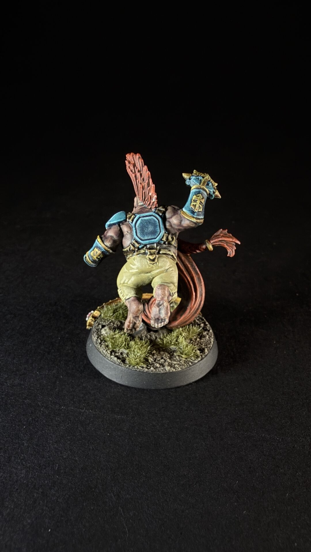 Back view of a painted fantasy miniature wearing blue armor and yellow shorts, with a red tail and a grassy rocky base.