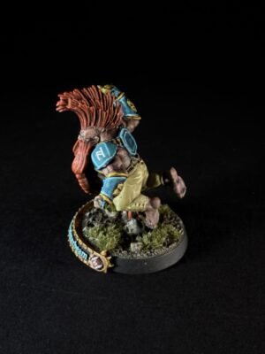 Painted tabletop miniature of a muscular humanoid with a red spiky mane, blue armor plates, and tan clothing, mid-stride on a rocky, grassy base.