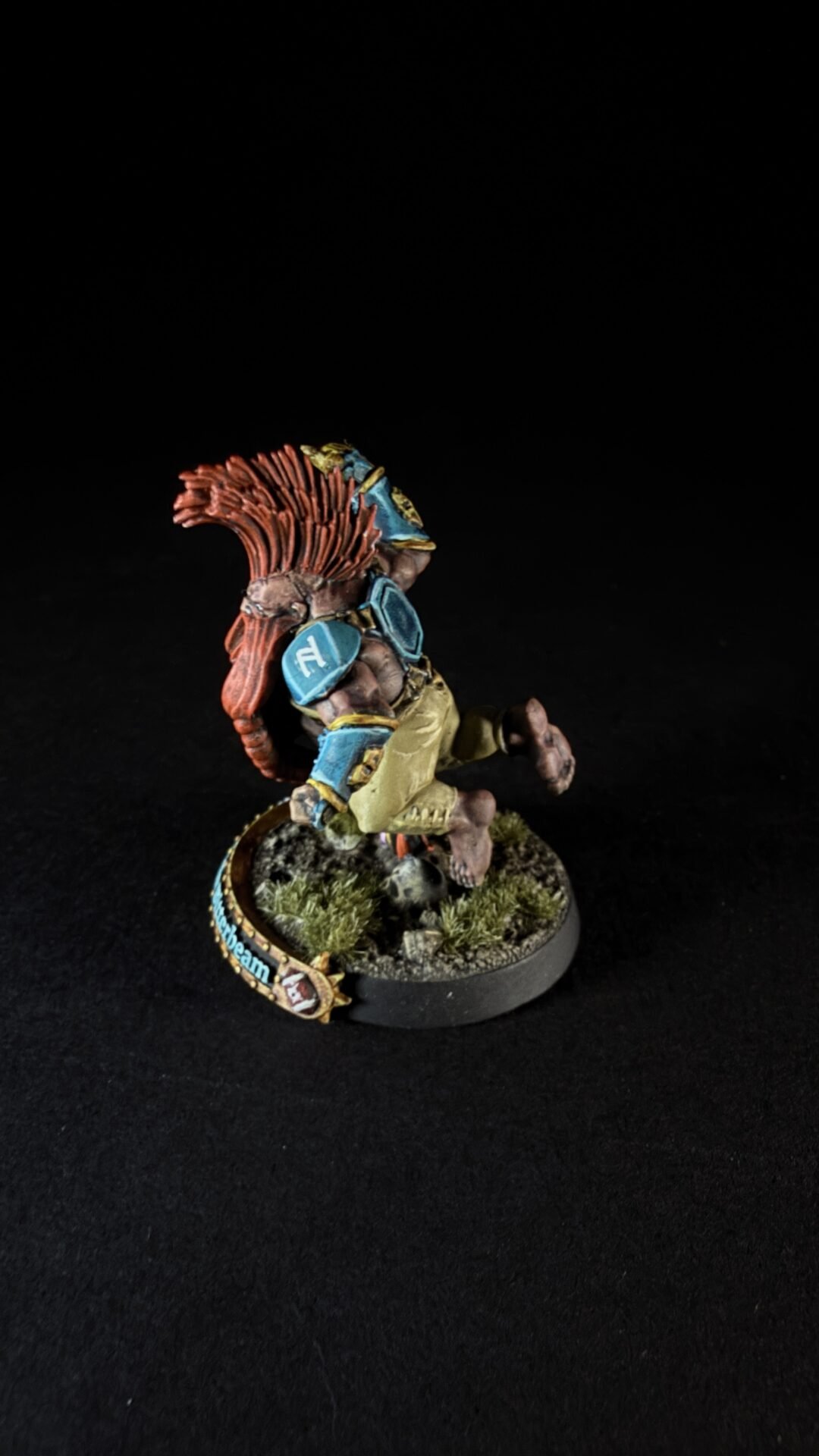 Painted tabletop miniature of a muscular humanoid with a red spiky mane, blue armor plates, and tan clothing, mid-stride on a rocky, grassy base.