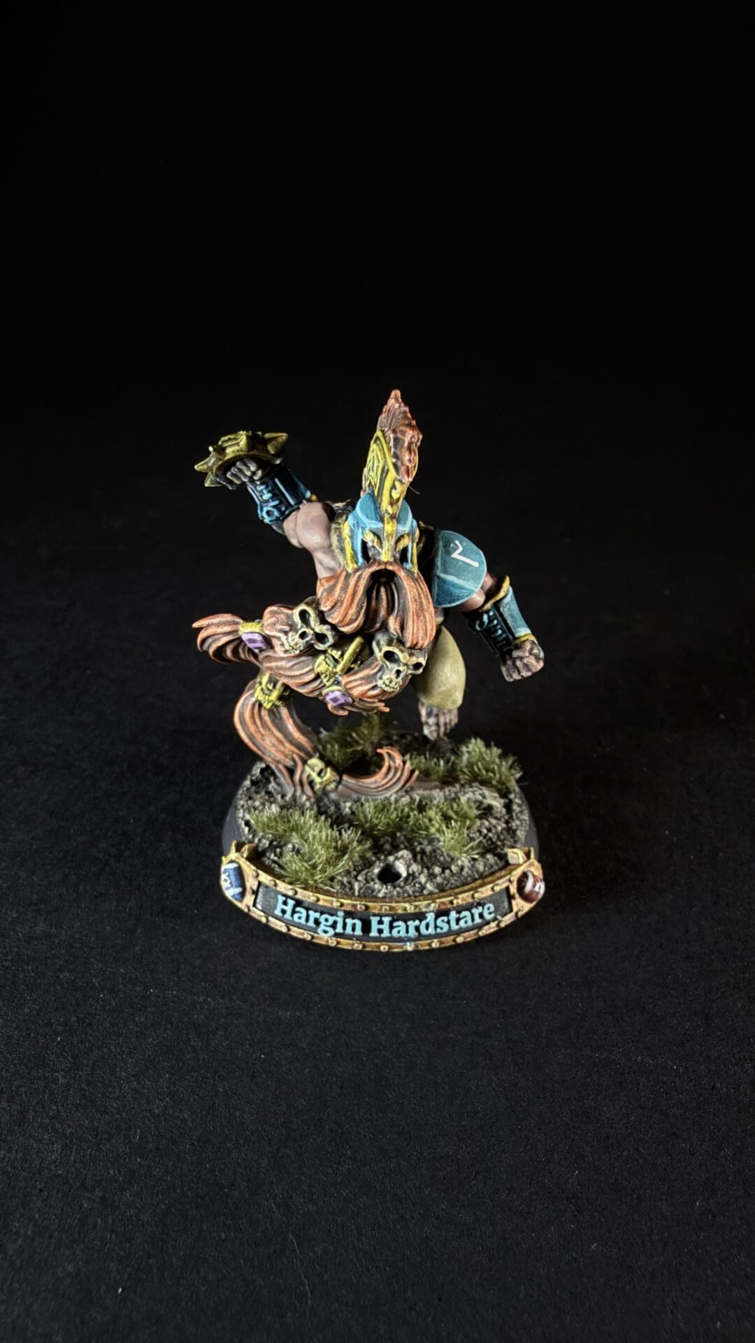 Painted tabletop miniature of a bearded dwarf in teal, yellow, and blue armor, posed on a rocky base with grass; nameplate reads 'Hargin Hardstare'