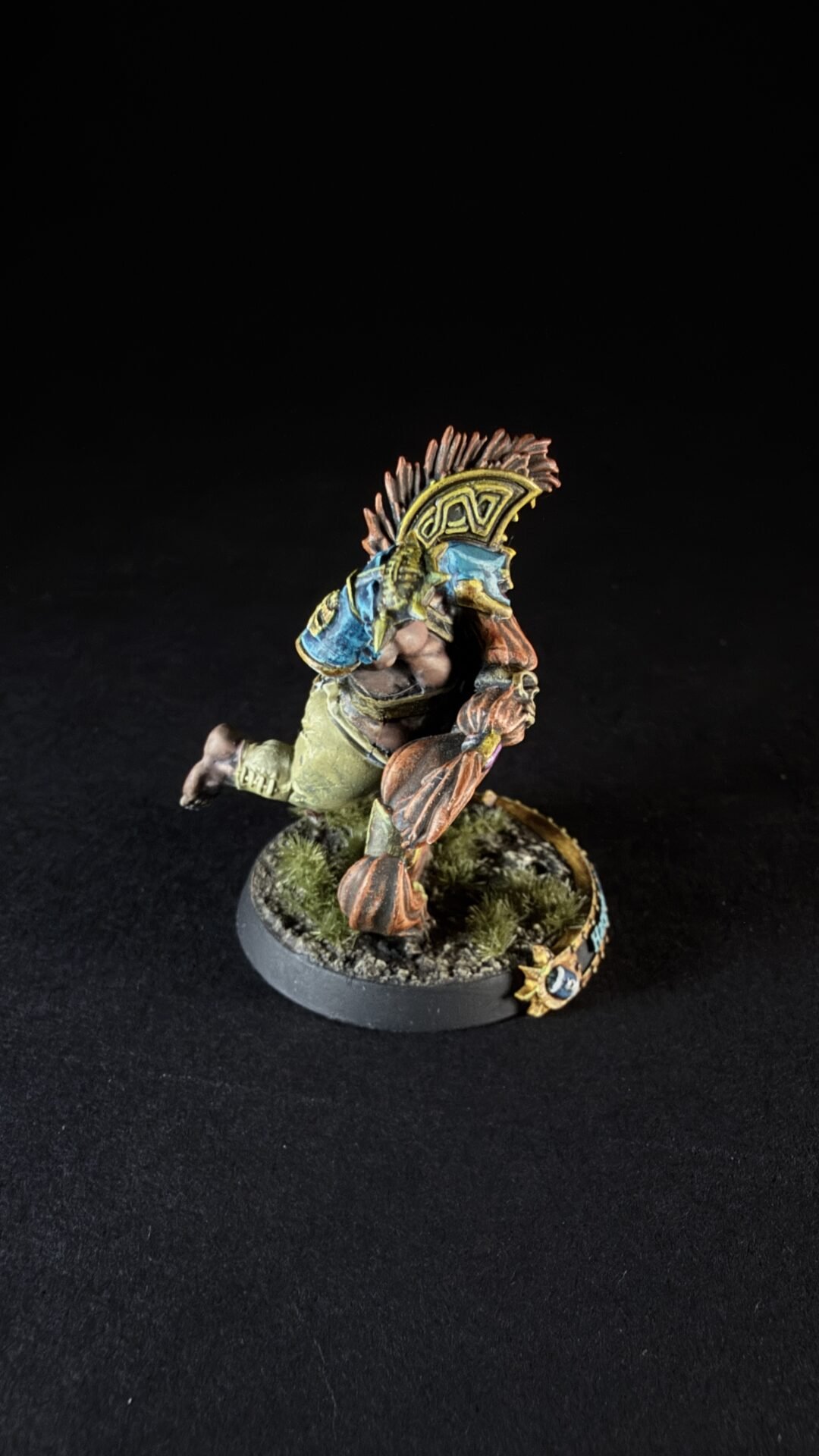 Tabletop miniature of a muscular orc/barbarian-wearing blue armor, mid-stride on a grassy base, with a decorative shield on its back.
