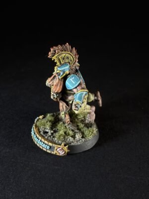 Wargaming miniature of a tribal warrior or orc with a feathered headdress, blue armor plates, and a weapon, posed on a rocky grassy base with a decorative banner on the edge (text visible).