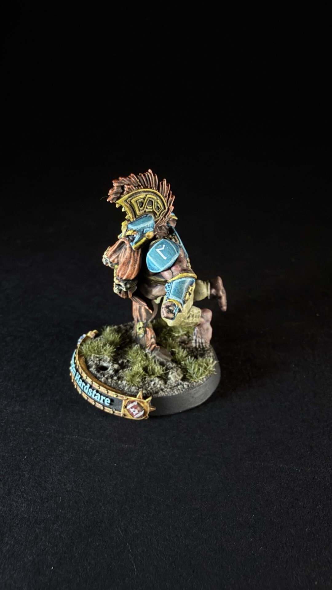 Wargaming miniature of a tribal warrior or orc with a feathered headdress, blue armor plates, and a weapon, posed on a rocky grassy base with a decorative banner on the edge (text visible).