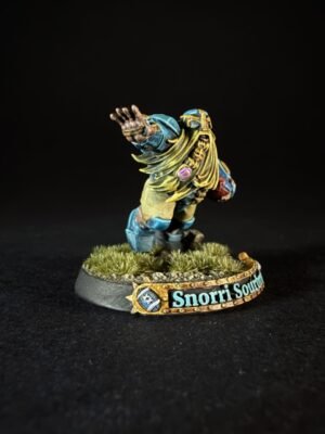 Fantasy tabletop miniature: a bearded warrior in blue and gold armor raises his right arm, standing on a grassy base with a decorative banner reading 'Snorri South...'.