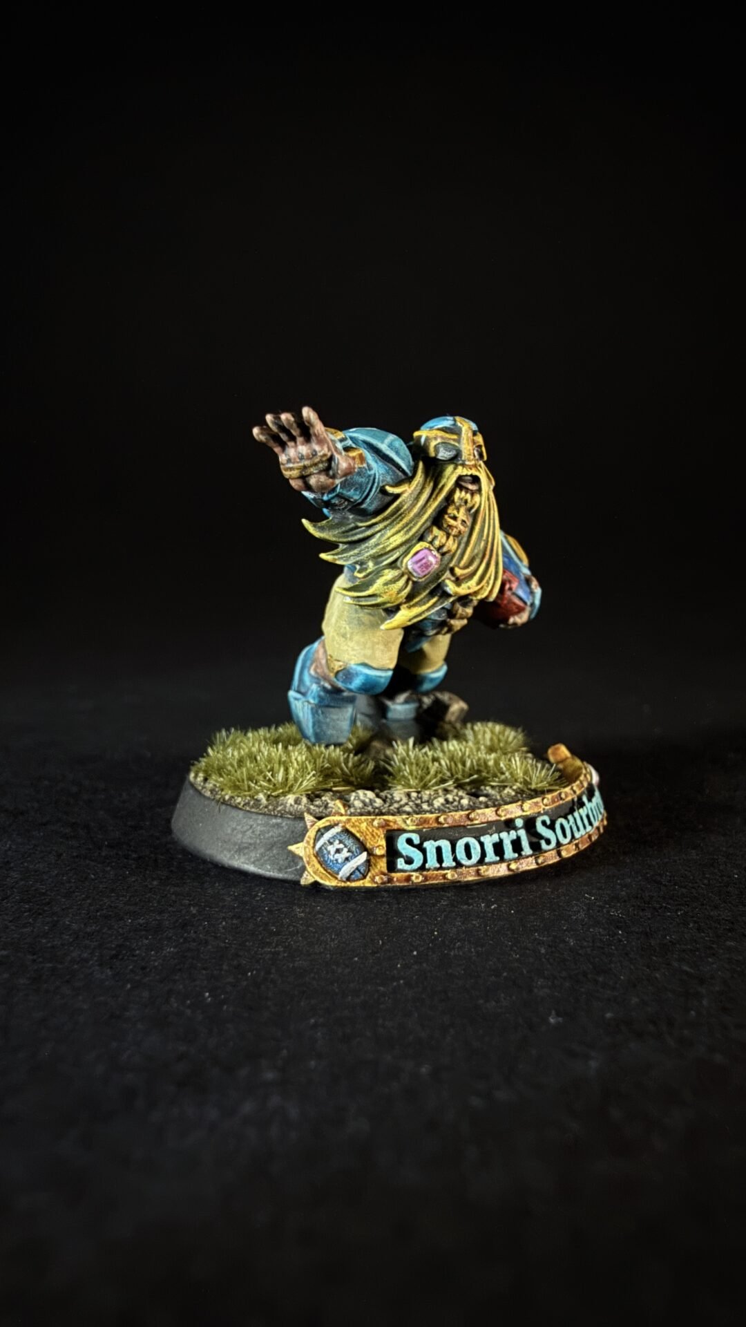 Fantasy tabletop miniature: a bearded warrior in blue and gold armor raises his right arm, standing on a grassy base with a decorative banner reading 'Snorri South...'.