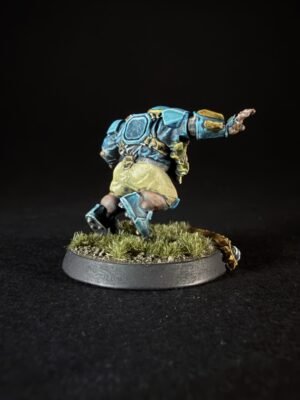 Miniature sci-fi soldier in blue armor leaping forward on a grassy base, viewed from the side against a dark backdrop.